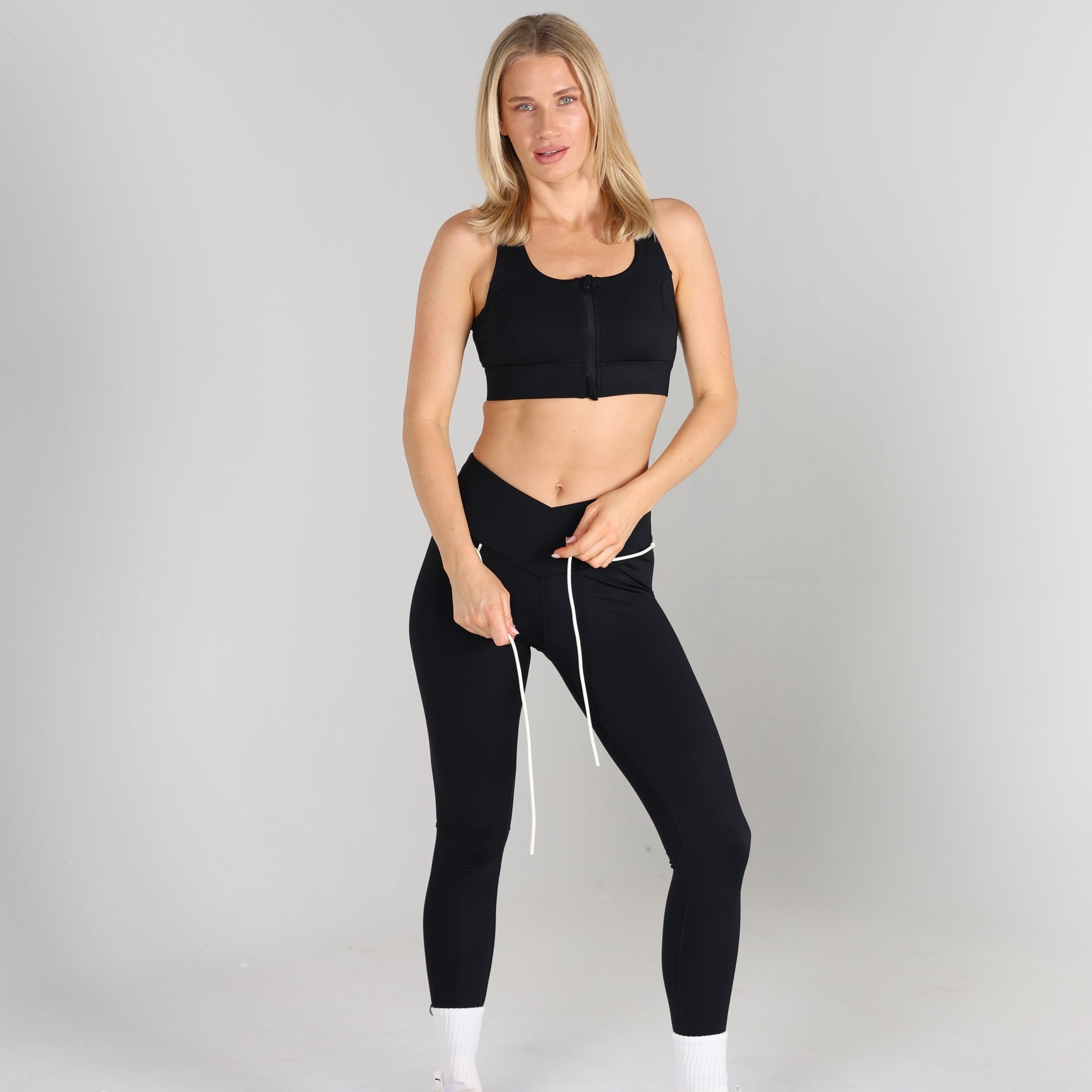 Woman standing, wearing a black zip-front cropped sports bra and black high-waisted leggings with white drawstring ties, white sneakers and socks, studio background.