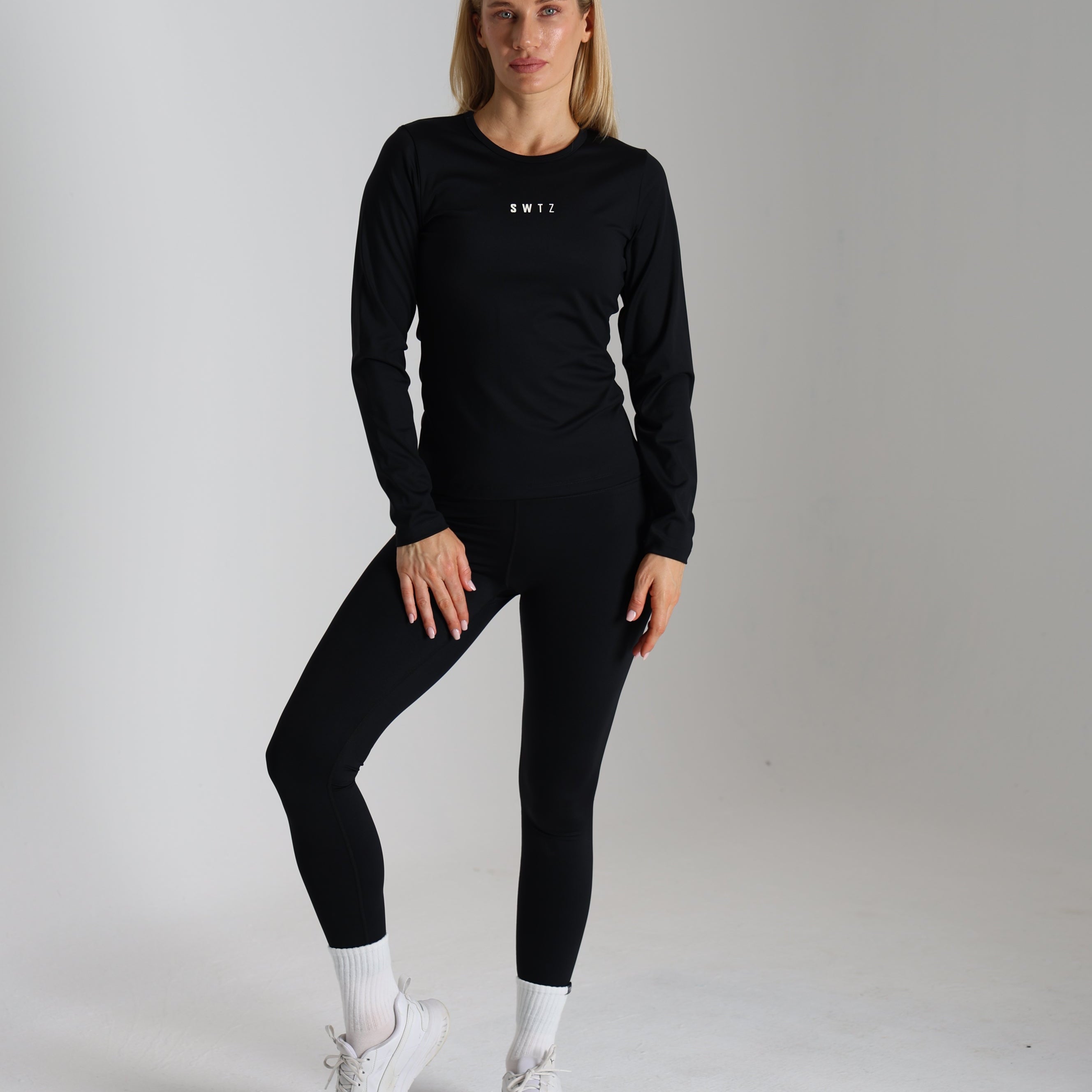 Woman standing, wearing a black long-sleeve top with SWTZ logo and black leggings, white sneakers and socks, studio background.